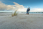 Zwei Personen gehen am Ostseestrand im Sand spazieren. Im Hintergrund ist das Meer, der Himmel ist blau mit weißen Wolken, im Vordergrund ist Schilf zu sehen. Die Personen tragen Mützen und dicke Jacken und gehen dicht beieinander. 