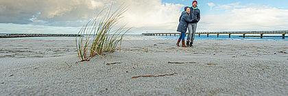 Zwei Personen spazieren am Ostseestrand. Sie tragen Mützen und dicke Jacken, das Meer ist im Hintergrund zu sehen, Schilf im Vordergrund. Der Himmel ist blau mit einigen weißen Wolken.  - zum Artikel 'Buchungslage im Schleswig-Holstein Tourismus für Herbst und Winter/Jahreswechsel '