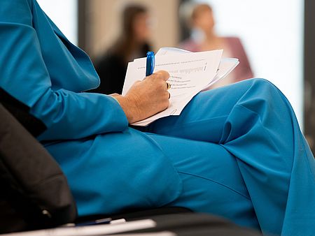 Eine Person in einem auffälligen, leuchtend blauen Anzug sitzt mit überschlagenen Beinen und schreibt mit einem blauen Kugelschreiber auf einem Papierstapel. 