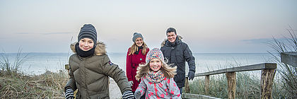 Eine Familie macht einen Strandspaziergang im Winter in Scharbeutz an der Ostsee. Frau, Mann und zwei Kinder laufen im Sand der Kamera entgegen. Im Hintergrund befindet sich das Meer, rechts und links Schilf und Dünenbefestigung aus Holz.  - zum Artikel 'Buchungslage im Schleswig-Holstein Tourismus für Weihnachten und den Jahreswechsel mit einem Ausblick auf das Frühjahr 2024'