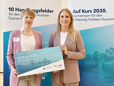 Britta Franke und Staatssekretärin Julia Carstens stehen nebeneinander vor einer blau gestalteten Informationswand mit der Überschrift „Auf Kurs 2030. Gemeinsam für den Schleswig-Holstein-Tourismus“. Beide blicken in die Kamera und lächeln. Sie halten gemeinsam ein großes, querformatiges Schild mit der Aufschrift „Auf Kurs 2030“.