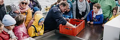 Eine Gruppe von Kindern und Erwachsenen steht dicht gedrängt auf einem Forschungsschiff an Deck. Sie beobachten aufmerksam einen Mann in dunkelblauer Kleidung, der aus einer roten Kiste Meereslebewesen entnimmt und zeigt. - zum Artikel 'Kurze Auszeiten: Ein Tag im echten Norden'