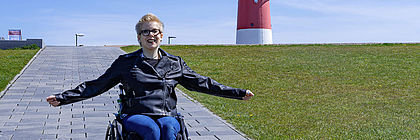 Eine Rollstuhlfahrerin befindet sich auf einem Deich. Im Hintergrund befindet sich der rot-weiße Leuchtturm von Büsum. - zum Artikel 'Barrierefrei/-arm durch den echten Norden'