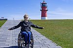 Eine Rollstuhlfahrerin befindet sich auf einem Deich. Im Hintergrund befindet sich der rot-weiße Leuchtturm von Büsum.