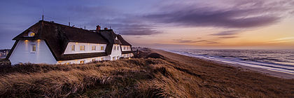 Ein großes, weißes Reetdachhaus steht auf einer Düne auf Sylt. Die Abenddämmerung hat eingesetzt. - zum Artikel 'Außergewöhnlich schlemmen in Schleswig-Holstein'