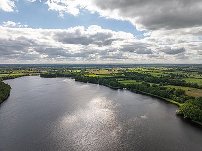 Luftaufnahme eines von Bäumen gesäumten Sees, umgeben von grünen Feldern und Wiesen unter einem weitläufigen, wolkenreichen Himmel.
