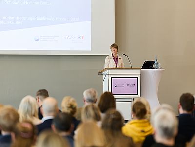 Britta Franke steht an einem Rednerpult in einem hellen Veranstaltungssaal und hält einen Vortrag vor einem sitzenden Publikum. Sie trägt einen hellen Blazer und spricht in ein Mikrofon. Hinter ihr ist eine Präsentationsfolie auf eine große Leinwand projiziert.