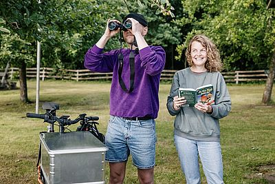 Zwei Personen beobachten Vögel im Grünen – eine mit Fernglas, die andere mit Bestimmungsbuch „Was fliegt denn da?“. Daneben steht ein Fahrrad.