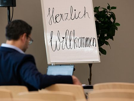 Ein Flipchart mit der handschriftlichen Aufschrift „Herzlich Willkommen“ steht vorne im Raum. Die Worte sind in großer, dekorativer Schrift geschrieben, wobei ein Herzsymbol als Punkt über dem „i“ im Wort „Willkommen“ verwendet wurde.