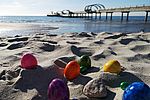 Bunte Ostereier liegen im Sand am Strand. Im Hintergrund befindet sich eine Seebrücke.