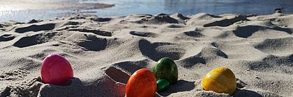 Bunte Ostereier liegen im Sand am Strand. Im Hintergrund befindet sich eine Seebrücke. - zum Artikel 'Frohe Ostern in Schleswig-Holstein!'
