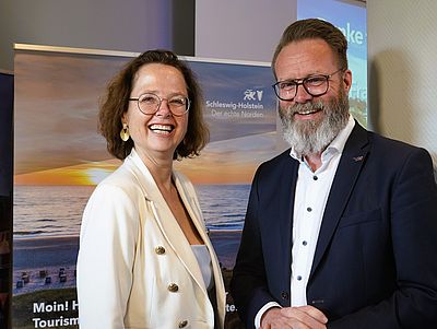 Dr. Bettina Bunge und Claus Ruhe Madsen stehen nebeneinander vor einem Aufsteller mit der Aufschrift „Schleswig-Holstein“ und einem Bild von Meer und Sonnenuntergang.
