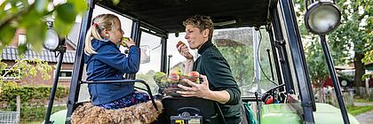 Eine Frau und ein Mädchen sitzen gemeinsam auf einem grünen Traktor. Das Mädchen trägt eine blaue Jacke und isst einen Apfel, während sie auf einem Fellpolster sitzt. Die Frau, in einem dunkelgrünen Pullover, hält einen Korb voller Äpfel auf dem Schoß und probiert ebenfalls einen. - zum Artikel 'Kohlanschnitt, Teezeit und Krabbentage'