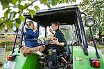 Eine Frau und ein Mädchen sitzen gemeinsam auf einem grünen Traktor. Das Mädchen trägt eine blaue Jacke und isst einen Apfel, während sie auf einem Fellpolster sitzt. Die Frau, in einem dunkelgrünen Pullover, hält einen Korb voller Äpfel auf dem Schoß und probiert ebenfalls einen.