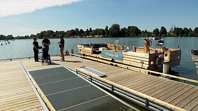 Barrierefreier Steg mit Rollstuhlfahrerin und Begleitung vor einem Floß auf einem ruhigen See bei sonnigem Wetter.