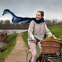 Eine lachende Frau fährt auf einem Fahrrad mit einem großen Weidenkorb vorn durch eine grüne, ländliche Landschaft. Der schmale Weg führt an einem Gewässer entlang. Die Frau trägt einen beigen Pullover, eine helle Hose und einen dunkelblauen Schal, der im Wind flattert.
