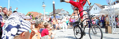 Ein Straßenkünstler macht Kunststücke auf einem abstrakten Fahrrad. Erwachsene und Kinder betrachten die Show. - zum Artikel 'Feste, Konzerte & Sport-Events: Kultur pur in Schleswig-Holstein'
