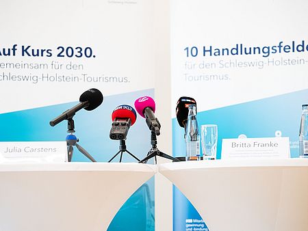 Zwei mit weißen Tischtüchern bedeckte Stehtische stehen nebeneinander, vorbereitet für eine Pressekonferenz oder Podiumsdiskussion. Auf den Tischen stehen Namensschilder mit den Aufschriften „Julia Carstens“ und „Britta Franke“. Vor den Schildern sind mehrere Mikrofone mit Logos verschiedener Medien.