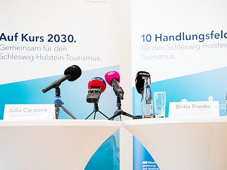 Zwei mit weißen Tischtüchern bedeckte Stehtische stehen nebeneinander, vorbereitet für eine Pressekonferenz oder Podiumsdiskussion. Auf den Tischen stehen Namensschilder mit den Aufschriften „Julia Carstens“ und „Britta Franke“. Vor den Schildern sind mehrere Mikrofone mit Logos verschiedener Medien.