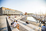 Menschen sitzen in Sommerkleidung auf großen Stufenterrassen am Segelhafen, der zum Ferienresort BeachBay auf dem Priwall in Lübeck-Travemünde gehört. Im Hintergrund sieht man das Hotel SlowDown, in dem es Workation-Angebote gibt. Das Wetter ist sommerlich. 