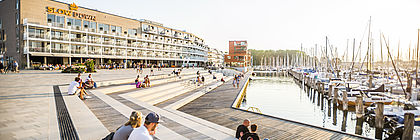 Menschen sitzen in Sommerkleidung auf großen Stufenterrassen am Segelhafen, der zum Ferienresort BeachBay auf dem Priwall in Lübeck-Travemünde gehört. Im Hintergrund sieht man das Hotel SlowDown, in dem es Workation-Angebote gibt. Das Wetter ist sommerlich.  - zum Artikel 'Arbeiten, wo andere Urlaub machen: Workation in Schleswig-Holstein '