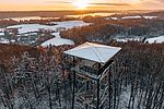 Im Vordergrund der Holzbergturm in Malente. Im Hintergrund der Blick über die wunderschöne Winterlandschaft der Holsteinischen Schweiz. Der Aussichtsturm ermöglicht einen Panoramablick. Die Wälder und Wiesen sind leicht schneebedeckt. Die Sonne scheint. 