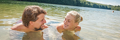 Ein Mann und eine Frau liegen in Badekleidung in einem See und lächeln sich an. Sie stützen sich mit den Unterarmen auf dem Boden ab. - zum Artikel 'Daten sind das Gold des digitalen Zeitalters - Touristische Landesdatenbank Schleswig-Holstein'