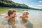 Ein Mann und eine Frau liegen in Badekleidung in einem See und lächeln sich an. Sie stützen sich mit den Unterarmen auf dem Boden ab.
