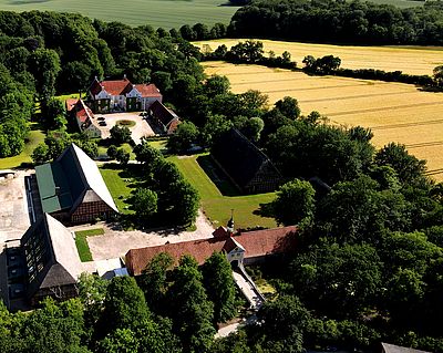 Luftaufnahme eines historischen Gutshofs mit mehreren Gebäuden, Wiesen und umliegenden Feldern am Waldrand.