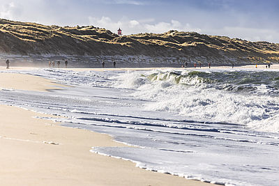 Urlauber spazieren am Strand des Sylter Ellenbogens bei tosender Nordseebrandung und strahlendem Herbstsonnenschein. Im Hintergrund ragt die Spitze des Leuchtfeuers List-West hervor.