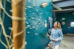 Eine Frau mit langen blonden Haaren und hellblauer Jeansjacke steht in einer maritim gestalteten Ausstellung. Sie blickt lächelnd auf eine Wand mit zahlreichen modellierten Fischen, die in Schwärmen angebracht sind.