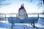 Eine Frau sitzt auf der Rückenlehne einer Bank und hat die Füße auf der Sitzfläche abgestellt. Sie blickt auf den verschneiten See.