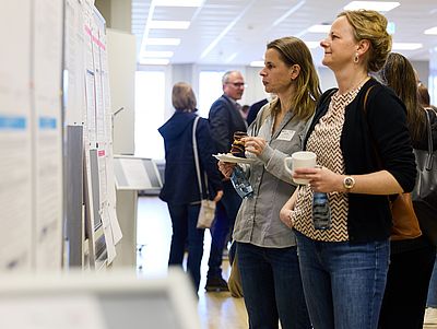 Zwei Frauen stehen in einem hellen Flur vor Stellwänden und lesen ausgehängte Informationen. Eine hält eine Tasse Kaffee und eine Wasserflasche, die andere einen Teller mit Gebäck und ebenfalls eine Flasche.