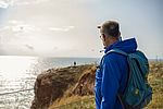 Ein Mann mit blauer Jacke und Rucksack blickt über eine Klippe hinweg auf das Meer. Zum Schutz vor der blendenden Sonne hält er eine Hand vors Gesicht.