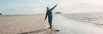 Eine Person in Winterkleidung läuft am Strand entlang und breitet die Arme aus. - zum Artikel 'Outdoor-Ausflugstipps gegen den Winterblues'