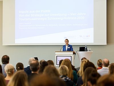 Staatssekretärin Julia Carstens steht an einem Rednerpult in einem großen, hellen Veranstaltungssaal und spricht vor einem sitzenden Publikum. Sie trägt einen blauen Blazer und spricht in ein Mikrofon. Neben ihr stehen ein Laptop sowie Wasserflaschen und Gläser auf einem Stehtisch. Hinter ihr ist eine große Präsentationsfolie mit dem Titel zur Tourismusstrategie Schleswig-Holstein 2030 projiziert.