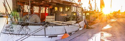 Der urige Fischereihafen in Travemünde wird vom Sonnenuntergang in ein goldenes Licht getaucht.  - zum Artikel 'Schleswig-Holstein feiert den Fisch!'