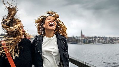 Zwei junge Frauen stehen draußen im Wind, ihre Haare werden stark verweht. Beide lachen ausgelassen. Die Frau links trägt dunkle Kleidung und hat blondes, offenes Haar. Die Frau rechts hat lockiges braunes Haar, trägt eine schwarze Lederjacke und ein weißes T-Shirt. 