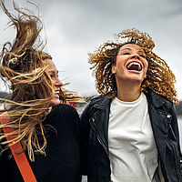 Zwei junge Frauen stehen draußen im Wind, ihre Haare werden stark verweht. Beide lachen ausgelassen. Die Frau links trägt dunkle Kleidung und hat blondes, offenes Haar. Die Frau rechts hat lockiges braunes Haar, trägt eine schwarze Lederjacke und ein weißes T-Shirt. 