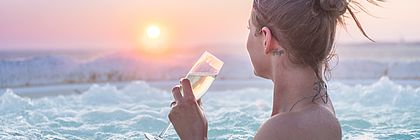 Eine Frau mit nackten Schultern sitzt in einem Außen-Whirlpool auf der Dachterrasse der Ostsee-Therme Scharbeutz und blickt vom Betrachter in Richtung Sonnenuntergang auf der Ostsee. In den Händen hält sie ein Glas Sekt. Man sieht das blubbernde Wasser des Whirlpools, den leicht schneebedeckten Deich und die Ostsee im Hintergrund.  - zum Artikel 'Karnevalsflucht: Es locken Wellnessoasen und unvergleichliche Naturerlebnisse in Schleswig-Holstein'