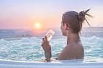 Eine Frau mit nackten Schultern sitzt in einem Außen-Whirlpool auf der Dachterrasse der Ostsee-Therme Scharbeutz und blickt vom Betrachter in Richtung Sonnenuntergang auf der Ostsee. In den Händen hält sie ein Glas Sekt. Man sieht das blubbernde Wasser des Whirlpools, den leicht schneebedeckten Deich und die Ostsee im Hintergrund. 