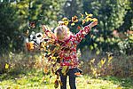 Ein blondes Kind in roter Jacke steht auf einer Wiese und wirft Herbstblätter in die Luft. Mit ausgebreiteten Armen blickt es nach unten, während die bunten Blätter um es herum durch die Luft wirbeln.