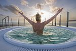 Eine Frau sitzt in einem Whirlpool, man sieht den Rücken und den Hinterkopf. Sie reckt die Arme gen Himmel. Ihr Blick geht von der Dachterrasse, wo sich der Whirlpool der Meerzeit Büsum befindet, über die Nordsee und in den Himmel über der See. Wasser spritzt. 