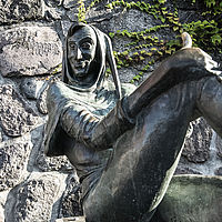 Bronzene Skulptur von Till Eulenspiegel vor einer groben Steinmauer. Die Figur trägt eine spitz zulaufende Kapuze und mittelalterliche Kleidung. Mit einem schelmischen Lächeln im Gesicht sitzt sie lässig, ein Bein angewinkelt und den Daumen hochgestreckt. 