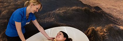 Eine Frau wird in einem Spa von einer Mitarbeiterin in eine Decke eingewickelt, beide lächeln.  - zum Artikel 'Von Kneipp bis Digital Detox: Gesund in den Herbst in  Schleswig-Holstein'