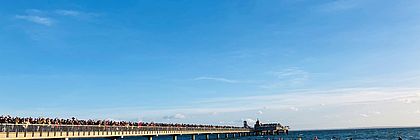 Eine große Gruppe von Menschen läuft vom Strand ins Meer, hier die Ostsee bei Grömitz. Alle tragen Badekleidung. Links auf der Seebrücke stehen Zuschauer und Zuschauerinnen, die das Winterbaden aus sicherer Entfernung verfolgen. Der Himmel ist strahlend blau.  - zum Artikel 'Aktiv durch den Winter – Outdoor-Erlebnisse in Schleswig-Holstein  '
