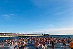 Eine große Gruppe von Menschen läuft vom Strand ins Meer, hier die Ostsee bei Grömitz. Alle tragen Badekleidung. Links auf der Seebrücke stehen Zuschauer und Zuschauerinnen, die das Winterbaden aus sicherer Entfernung verfolgen. Der Himmel ist strahlend blau. 