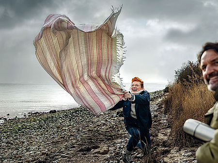 Eine fröhliche Frau mit rotem Haar steht auf einem steinigen Strandabschnitt und hält eine große, gestreifte Decke fest, die vom Wind aufgeweht wird. Sie lacht und wirkt ausgelassen. Rechts im Vordergrund lächelt ein Mann mit dunklen Haaren in die Kamera.