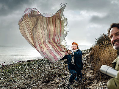 Eine fröhliche Frau mit rotem Haar steht auf einem steinigen Strandabschnitt und hält eine große, gestreifte Decke fest, die vom Wind aufgeweht wird. Sie lacht und wirkt ausgelassen. Rechts im Vordergrund lächelt ein Mann mit dunklen Haaren in die Kamera.