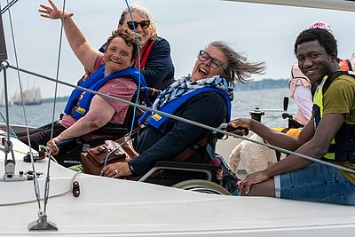 Vier Personen sitzen lachend in einem Segelboot, zwei davon in Rollstühlen. Alle tragen Schwimmwesten, der Wind weht durch die Haare.